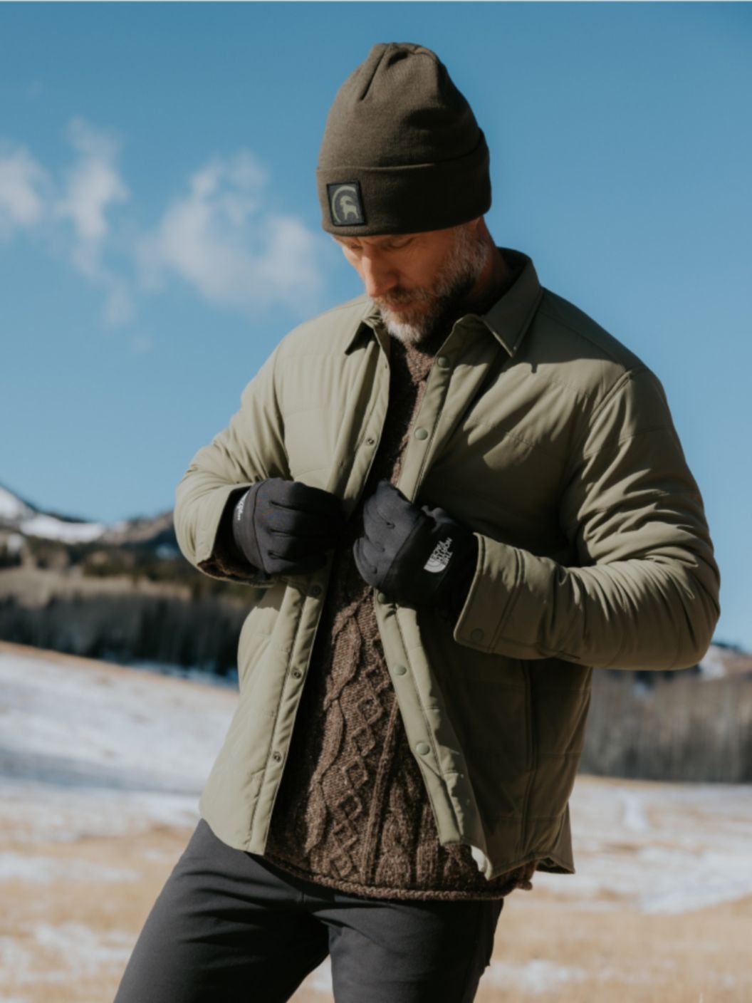 A man in a snowy field buttons an insulated jacket over a fisherman’s sweater.